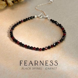May include: A delicate bracelet featuring black spinel and garnet beads. The beads are strung together with a silver chain and clasp. The word "FEARNESS" is printed below the bracelet, with "BLACK SPINEL • GARNET" underneath.