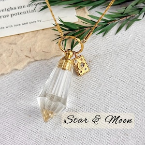 May include: A gold-colored necklace featuring a clear crystal pendant with a gold cap and a small rectangular charm with a moon and star design. The necklace is displayed on a white surface with green foliage.