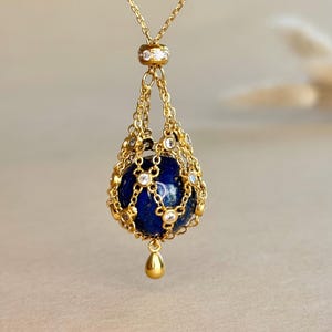 Peut inclure: Un pendentif collier doré composé d'une pierre sphérique en lapis-lazuli bleu profond enfermée dans une cage en fine chaîne dorée. La cage est ornée de petites pierres claires à chaque jonction, avec une breloque goutte dorée suspendue.