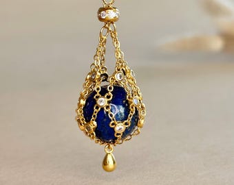 May include: A gold-toned pendant necklace featuring a spherical deep blue lapis lazuli stone encased in a delicate gold chain cage. The cage is accented with small clear stones at each junction, and a smooth gold teardrop charm hangs from the bottom.