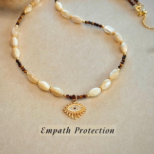 May include: A necklace with a gold-coloured eye pendant, set with crystals, and strung with white oval beads and brown beads. The necklace has gold-coloured accents. The text "Empath Protection" is below the necklace.