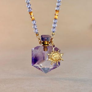 Puede incluir: Un collar con colgante de botella de perfume de cristal púrpura con un dije de sol dorado. El collar presenta una cadena de cuentas con detalles dorados. La botella tiene forma hexagonal facetada.