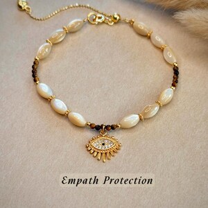May include: A gold bracelet with oval, cream-coloured beads and small brown beads. A gold charm with a sparkling eye design hangs from the centre. The text "Empath Protection" is at the bottom of the image.