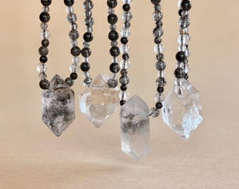 May include: Four necklaces featuring clear and black speckled crystal beads strung on thin cords. Each necklace terminates in a large, double-terminated hexagonal crystal pendant. The crystals vary in clarity, ranging from transparent to opaque black-flecked.