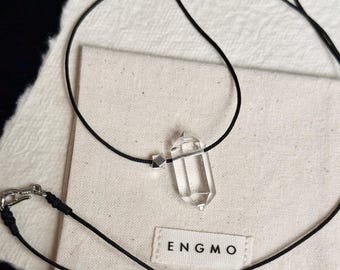 May include: A necklace featuring a clear, hexagonal, double-terminated crystal pendant suspended on a thin black cord. A small, metallic, geometric bead is positioned next to the crystal. The cord includes a silver-toned metal lobster clasp.
