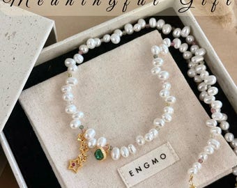 May include: A necklace made of white freshwater pearls interspersed with small multicoloured beads. The piece features a gold-toned charm with three star shapes and a single green stone. The text "Meaningful Gift" and "ENGMO" are visible.