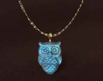 May include: A gold chain necklace featuring a carved owl pendant made of iridescent blue labradorite stone. The owl has detailed circular eyes and textured feather patterns on its body, suspended from a small gold bail with a tiny clear stone.