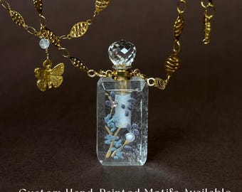 May include: A clear rectangular glass perfume bottle pendant with a faceted crystal stopper, featuring a hand-painted blue floral motif inside. The bottle hangs from a gold-toned chain with oval links and a small butterfly charm. Custom Hand-Painted Motifs Available.