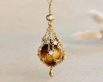 May include: A gold pendant necklace featuring a spherical tiger's eye gemstone encased in a delicate gold chain cage. The cage is accented with small, round, clear stones at each intersection. A small gold teardrop charm hangs from the bottom.
