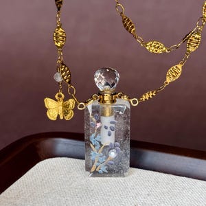 May include: A rectangular clear glass perfume bottle pendant featuring a floral design with blue and white accents, topped with a faceted crystal stopper. The pendant hangs from a gold-toned chain with textured oval links and a small butterfly charm.