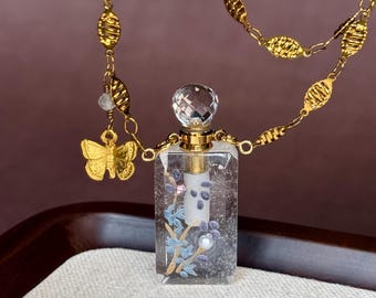 May include: A rectangular clear glass perfume bottle pendant featuring a floral design with blue and white accents, topped with a faceted crystal stopper. The pendant hangs from a gold-toned chain with textured oval links and a small butterfly charm.