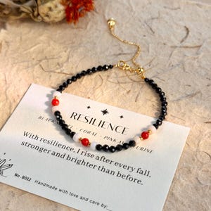 May include: A black beaded bracelet with red and white accents. The bracelet is on a card with the word "RESILIENCE" and the text "With resilience, I rise after every fall, stronger and brighter than before."