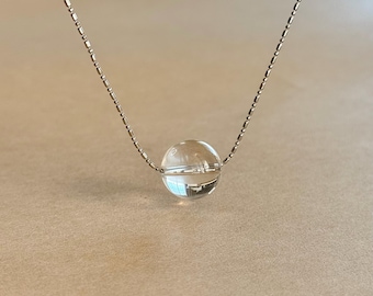 May include: A delicate necklace featuring a single, clear, spherical glass bead suspended on a thin, metallic, beaded chain. The transparent orb reflects light, creating a bright highlight on its surface, while the chain consists of small, uniform metal links.