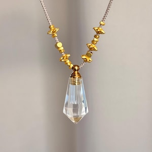 Puede incluir: Un collar con un colgante de cristal transparente de facetas geométricas suspendido de una tapa de metal dorado. La cadena consiste en un cordón gris claro con pequeñas cuentas doradas texturizadas y espaciadores en forma de estrella.