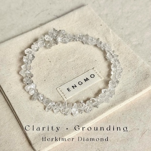 Puede incluir: Una pulsera circular hecha de pequeños cristales de diamante Herkimer transparentes y en bruto. La pulsera descansa sobre una bolsa de tela beige con el texto ENGMO, Clarity, Grounding y Herkimer Diamond impreso en tinta negra.