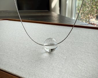 May include: A clear, spherical glass pendant suspended on a thin, metallic silver-toned chain. The transparent orb features a smooth, polished surface that reflects light, creating a simple and clean aesthetic for this piece of jewelry.