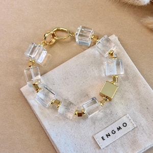 Puede incluir: Un brazalete dorado con cuentas transparentes en forma de cubo y conectores dorados. El brazalete descansa sobre una tela beige con una etiqueta "ENGMO". El diseño es moderno y elegante, perfecto para cualquier ocasión.