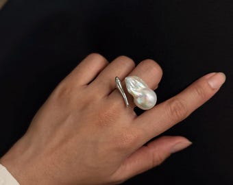 May include: A silver-toned open ring featuring a single large, irregular, iridescent white baroque pearl on one end and a smooth, tapered silver-toned metal point on the other end, creating a bypass style design on the finger.