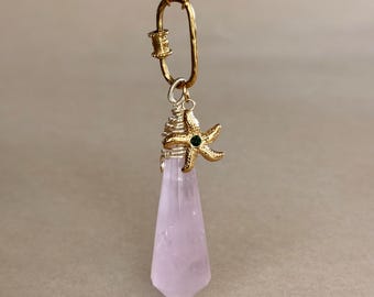 May include: A gold-colored chain necklace with a pendant featuring a light pink crystal point, a gold-colored starfish charm with a green gem, and a gold-colored clasp. The necklace is displayed against a neutral background.