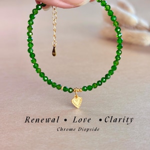May include: A delicate bracelet featuring faceted, vibrant green beads. A small, gold-colored heart charm dangles from the center. The text "Renewal • Love • Clarity" and "Chrome Diopside" are displayed below the bracelet.