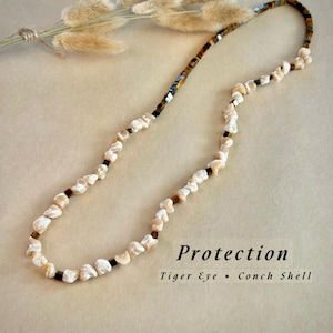 Op de afbeelding: Een ketting met schelp- en tijgeroogkralen. De ketting heeft een mix van witte, bruine en zwarte kralen. Het woord "Protection" staat in zwarte letters onderaan de afbeelding.
