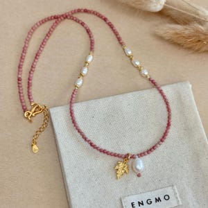 May include: A delicate necklace featuring small, pink beads and white pearls. A gold-colored leaf charm and a single pearl pendant hang from the necklace. The necklace has a gold-colored clasp. The necklace is displayed on a white cloth.