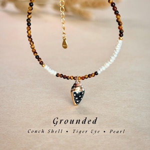 May include: A delicate necklace featuring a conch shell pendant with gold accents and black polka dots. The necklace is strung with tiger eye and pearl beads. The word "Grounded" is written below the necklace.