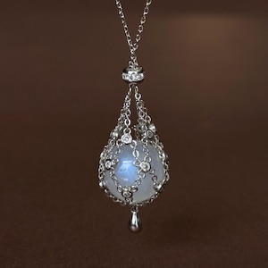 Puede incluir: Un collar con colgante de tono plateado que presenta una piedra preciosa esférica de color blanco translúcido, encerrada en una delicada red metálica adornada con pequeñas piedras redondas transparentes. Un pequeño dije de metal en forma de gota cuelga en la base.