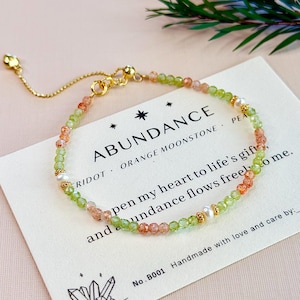 May include: A delicate bracelet featuring faceted green peridot and orange moonstone beads, accented with small white pearls and gold-toned metal spacers. The bracelet includes a gold-toned adjustable chain and rests on a white card with the text ABUNDANCE.