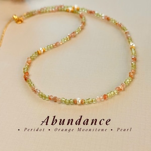 Puede incluir: Un collar de cuentas con peridoto verde facetado y piedra de luna naranja, acentuado con pequeñas perlas blancas y espaciadores dorados. El texto debajo dice: Abundance • Peridot • Orange Moonstone • Pearl.