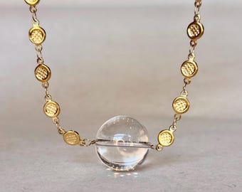 May include: A gold chain necklace featuring a series of small, circular gold links with a textured, grid-like pattern. A single, clear, spherical glass bead is suspended in the center of the chain, acting as a focal point for the jewelry piece.