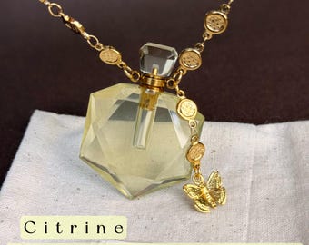 May include: A gold-toned necklace featuring a faceted, pale yellow, diamond-shaped pendant bottle with a clear stopper. The chain includes small circular accents and a gold butterfly charm. Text reads: Citrine - The Stone of Joy & Positivity.