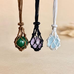 Può includere: Tre collane con pietre preziose sferiche tenute in reti di corda annodata. La collana a sinistra ha una sfera di malachite verde in corda marrone, quella centrale un'ametista viola chiaro in corda nera e quella a destra una sfera azzurra in corda bianca.