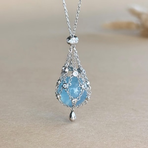Peut inclure: Un collier en argent avec un pendentif en forme de goutte d'eau bleu clair. Le pendentif est enfermé dans une cage en argent délicate ornée de petites pierres précieuses transparentes. Une petite goutte d'argent pend en dessous.