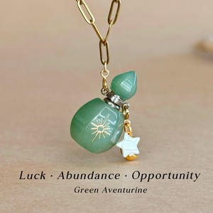 Może przedstawiać: Zielony naszyjnik z wisiorkiem z awenturynu ze złotym łańcuszkiem. Wisiorek ma kształt małej butelki ze wzorem słońca i zawieszką w kształcie gwiazdy. Tekst na dole brzmi "Luck • Abundance • Opportunity" i "Green Aventurine".