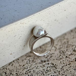 Può includere: Un anello in argento con una singola perla barocca bianca irregolare montata sulla parte superiore. La fascia è liscia e lucida, con un design aperto in cui il metallo si curva verso l'alto per sostenere la perla, creando un aspetto moderno.