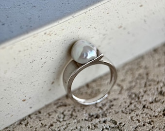 May include: A silver ring featuring a single, irregular, white baroque pearl set at the top. The band is smooth and polished, with a unique open-style design where the metal curves upward to cradle the pearl, creating a clean and modern aesthetic.
