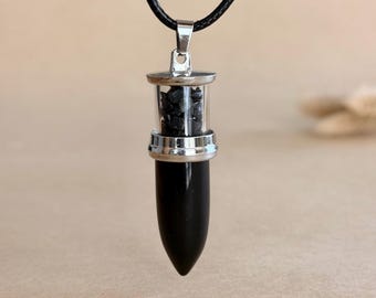 May include: A pendant necklace featuring a black cord and a cylindrical glass vial filled with small black stone chips, topped with a silver-toned metal cap and base, ending in a smooth, pointed black stone tip.