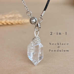 May include: A clear, double-terminated crystal pendant wrapped in silver-toned wire, attached to a silver chain and black cord. The text 2-in-1 Necklace & Pendulum is visible. The piece features a textured silver bead near the clasp.