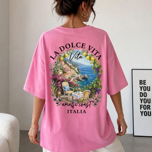 Peut inclure: Un t-shirt rose avec une impression graphique de la côte amalfitaine en Italie. Le motif comprend les mots "La Dolce Vita" et "Amalfi Coast Italia" avec des illustrations de citrons, de fleurs et d'une scène côtière.