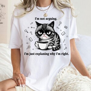 Peut inclure: T-shirt blanc avec un chat grincheux noir et gris tenant une tasse de café. Le texte sur le t-shirt dit : « Je ne discute pas. J'explique juste pourquoi j'ai raison. » Le chat a une expression renfrognée.