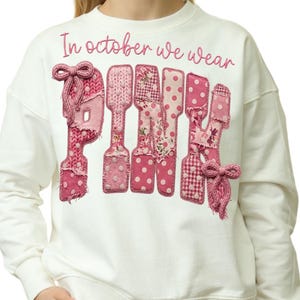 Peut inclure: Un sweat-shirt blanc à manches longues avec le texte In october we wear en écriture rose au-dessus du mot PINK. Les lettres sont composées de divers patchs en tissu rose avec des textures à pois, florales et tricotées, ornées de deux nœuds roses.