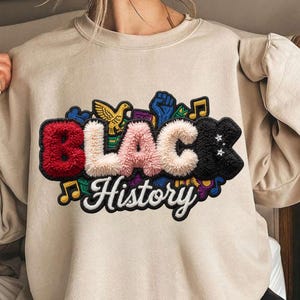 Peut inclure: Un sweat-shirt beige avec un écusson texturé indiquant "BLACK History". L'écusson comprend un "B" rouge, un "LAC" rose et un "K" noir avec des étoiles, entouré de notes de musique, d'un oiseau et d'un poing levé.