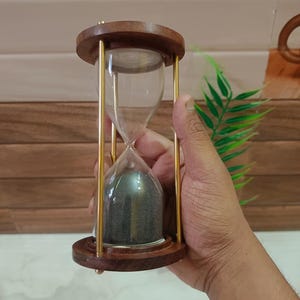May include: An hourglass with a wooden frame and base, featuring a clear glass chamber filled with dark sand. The hourglass is supported by gold-colored metal rods. The top and bottom are made of dark brown wood.
