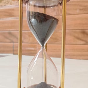 May include: An hourglass with a wooden frame and brass-coloured metal supports. The clear glass bulb contains black sand. The hourglass is a decorative item, often used as a timer or a symbol of time passing.