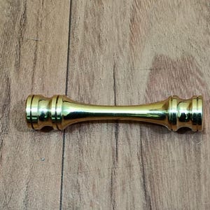 May include: A gold-colored, cylindrical furniture handle with a decorative design. The handle is approximately 4 inches long and has a symmetrical shape with rounded ends and a central, narrower section. It is set against a wooden background.