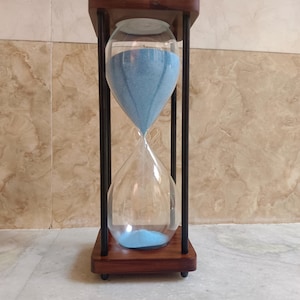 May include: An hourglass with a wooden frame and black supports. The clear glass bulb contains light blue sand. The hourglass is set on a wooden base.