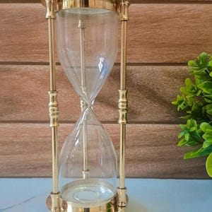 May include: A decorative hourglass with a clear glass body and a gold-colored metal frame. The hourglass has a classic design with a wide top and bottom, connected by slender metal rods. The sand timer is a timeless piece for home decor.