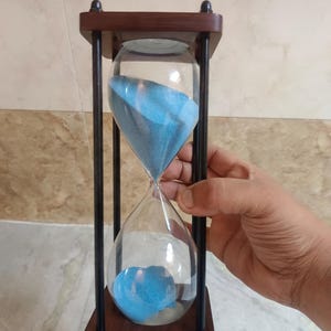 May include: An hourglass with a dark brown wooden frame and black vertical supports. The clear glass bulbs contain blue sand. The hourglass is held by a hand.