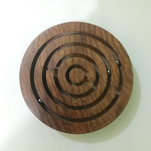 May include: A wooden labyrinth puzzle with a circular design. The puzzle features concentric circular grooves and several small metal balls within the maze. The wood has a rich, dark brown color.